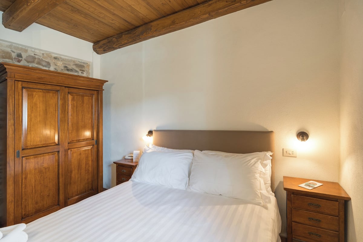 A bedroom with a bed, wooden wardrobe, bedside table, and a nightstand with a lamp.