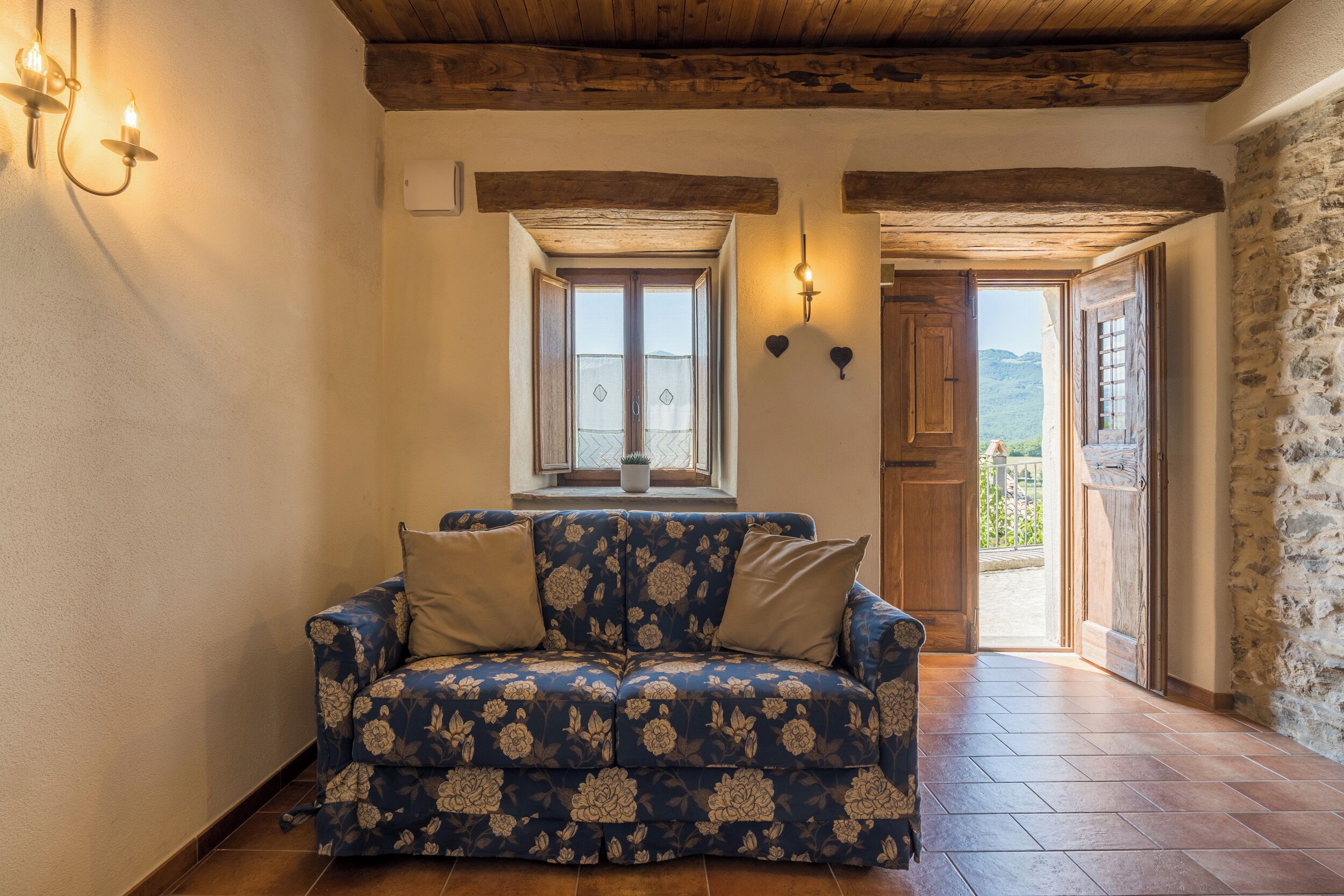 A cozy room with a floral-patterned sofa, a wooden ceiling, and a view of the outdoors through a door.