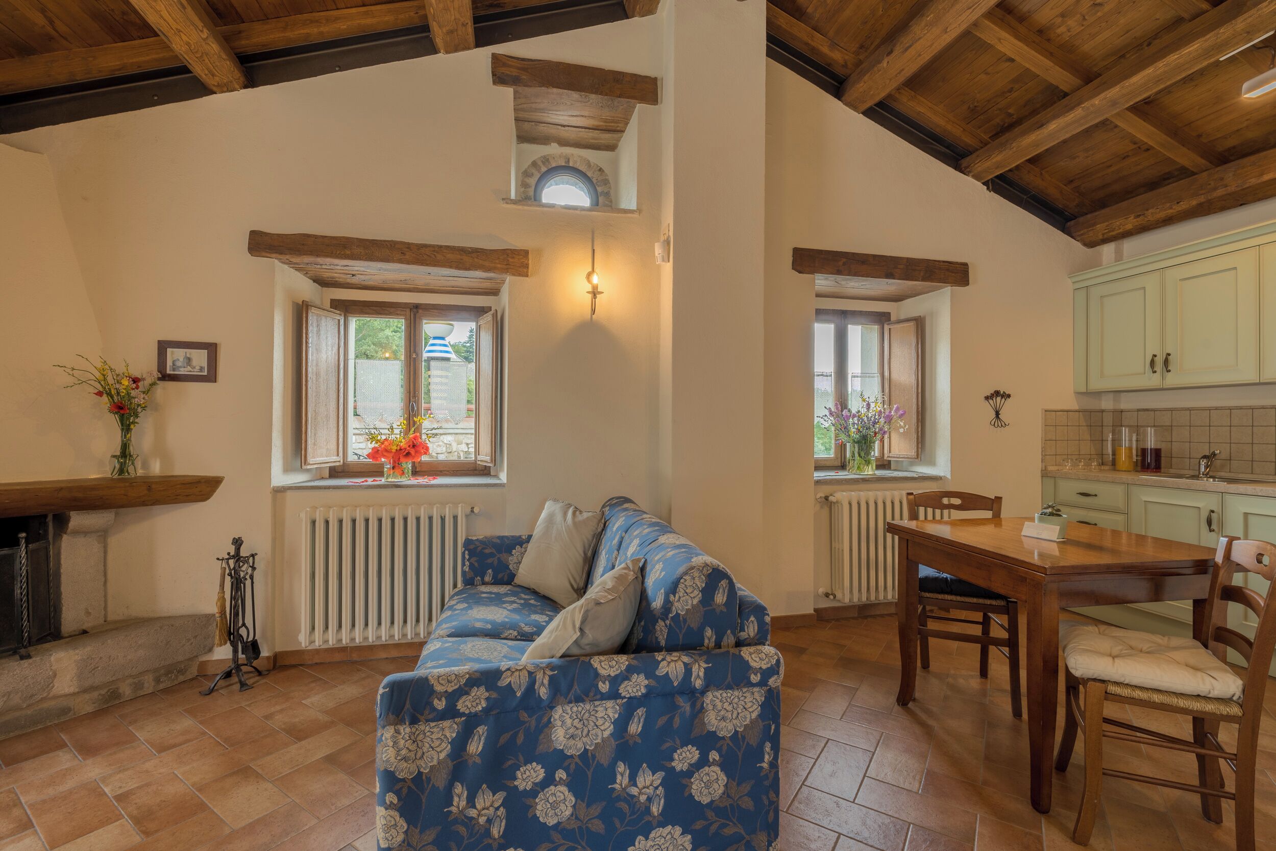 A cozy living room with a floral sofa, wooden dining table, and a kitchen area with light green cabinets.