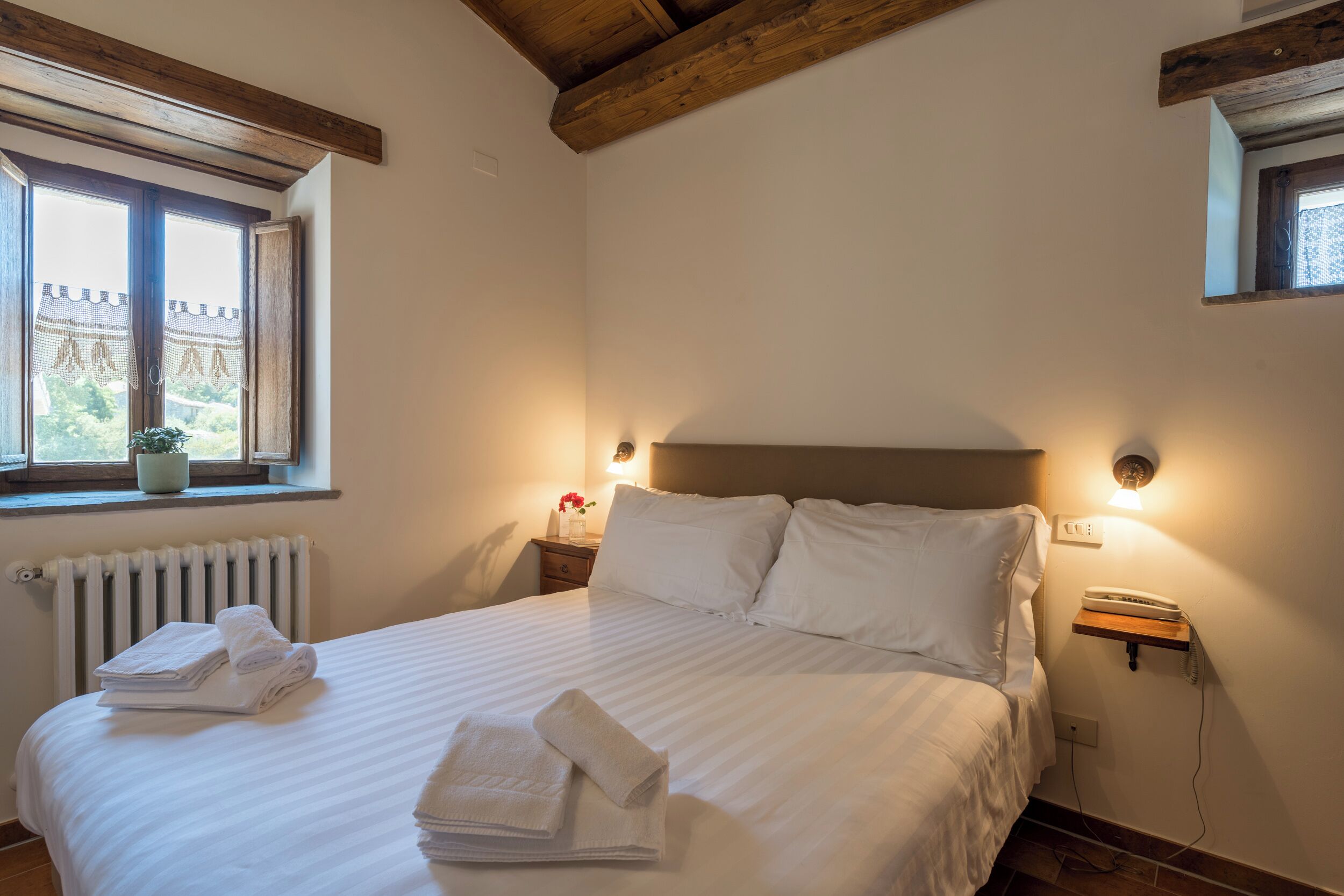 Cama bem arrumada com lençóis e toalhas brancas, mesa de cabeceira com abajur e janela com vista para o verde.
