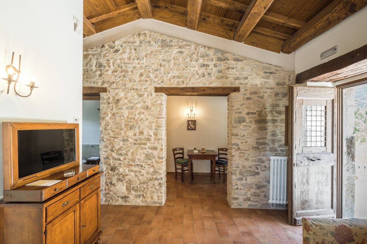 A rustic room with stone walls, wooden beams, a dining area with chairs, and a television on a wooden cabinet.