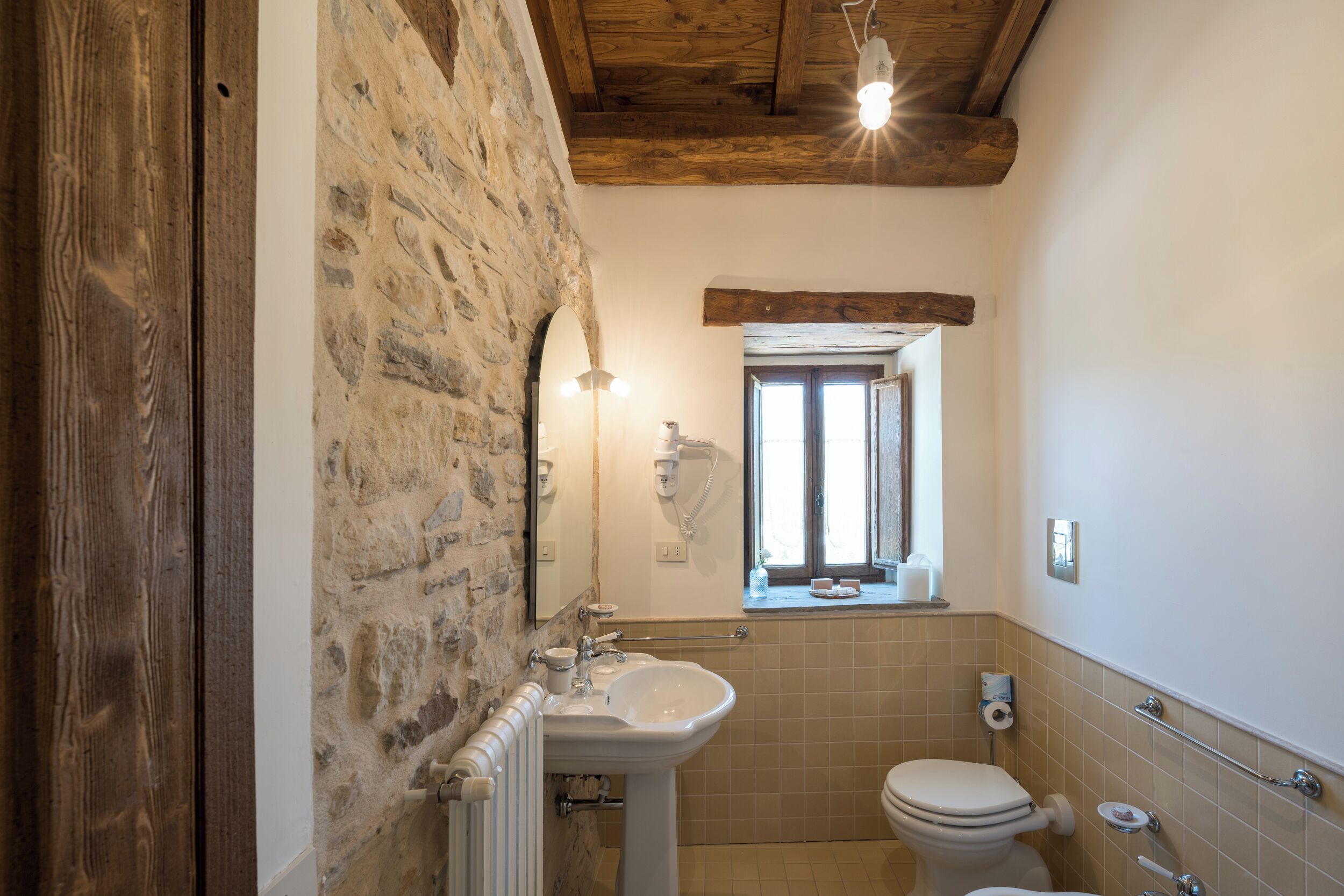 Une salle de bain avec un mur en pierre, des poutres en bois au plafond, un lavabo blanc et des toilettes.