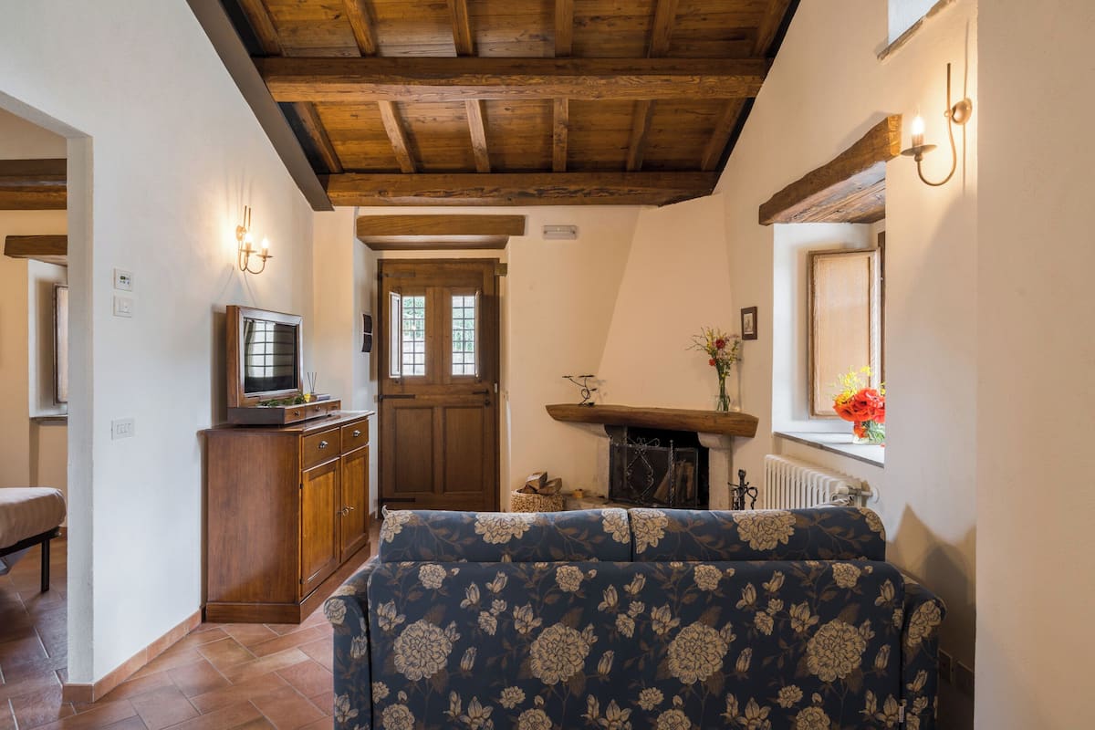 A cozy living room with a wooden ceiling, a television on a wooden cabinet, a floral sofa, and a window with flowers.