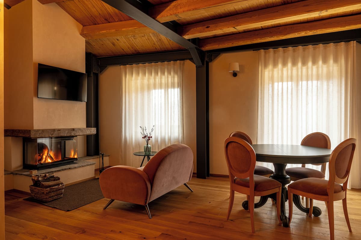 A cozy living room with a fireplace, wooden flooring, a round dining table with chairs, and a large window with sheer curtains.