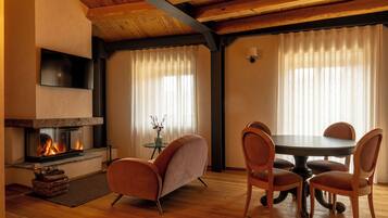 Un salon chaleureux avec une cheminée, un plancher en bois, une table à manger ronde entourée de chaises et une grande fenêtre ornée de rideaux légers.