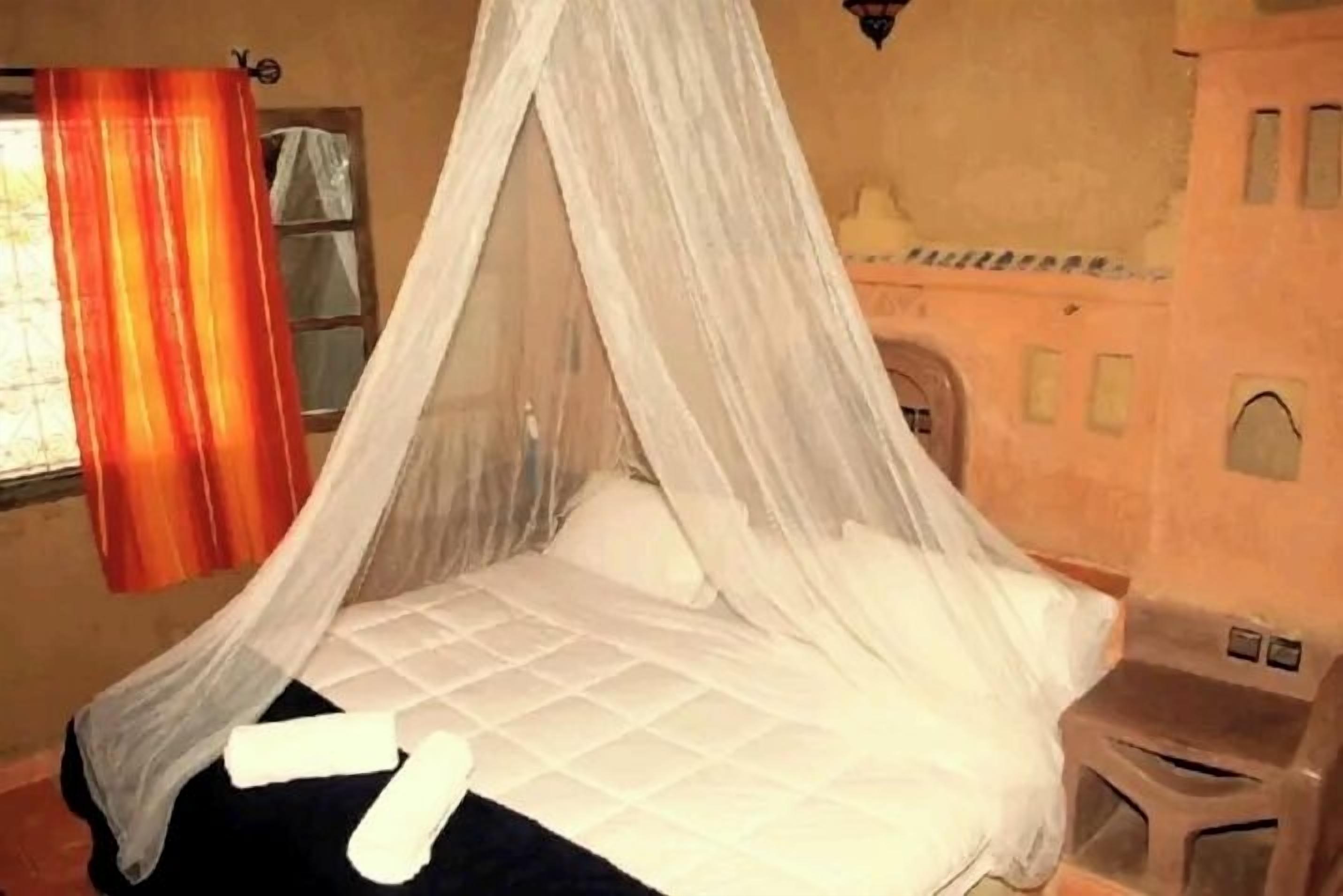 A room with a bed under a canopy, a window with curtains, and a small stool.