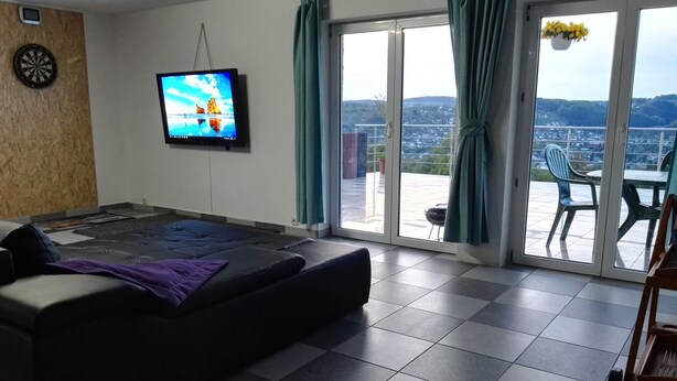 A living room with a sofa, a flat-screen TV, a sliding glass door leading to a balcony with outdoor furniture. - Ferienwohnungen nähe Badesee (Bergneustadt)