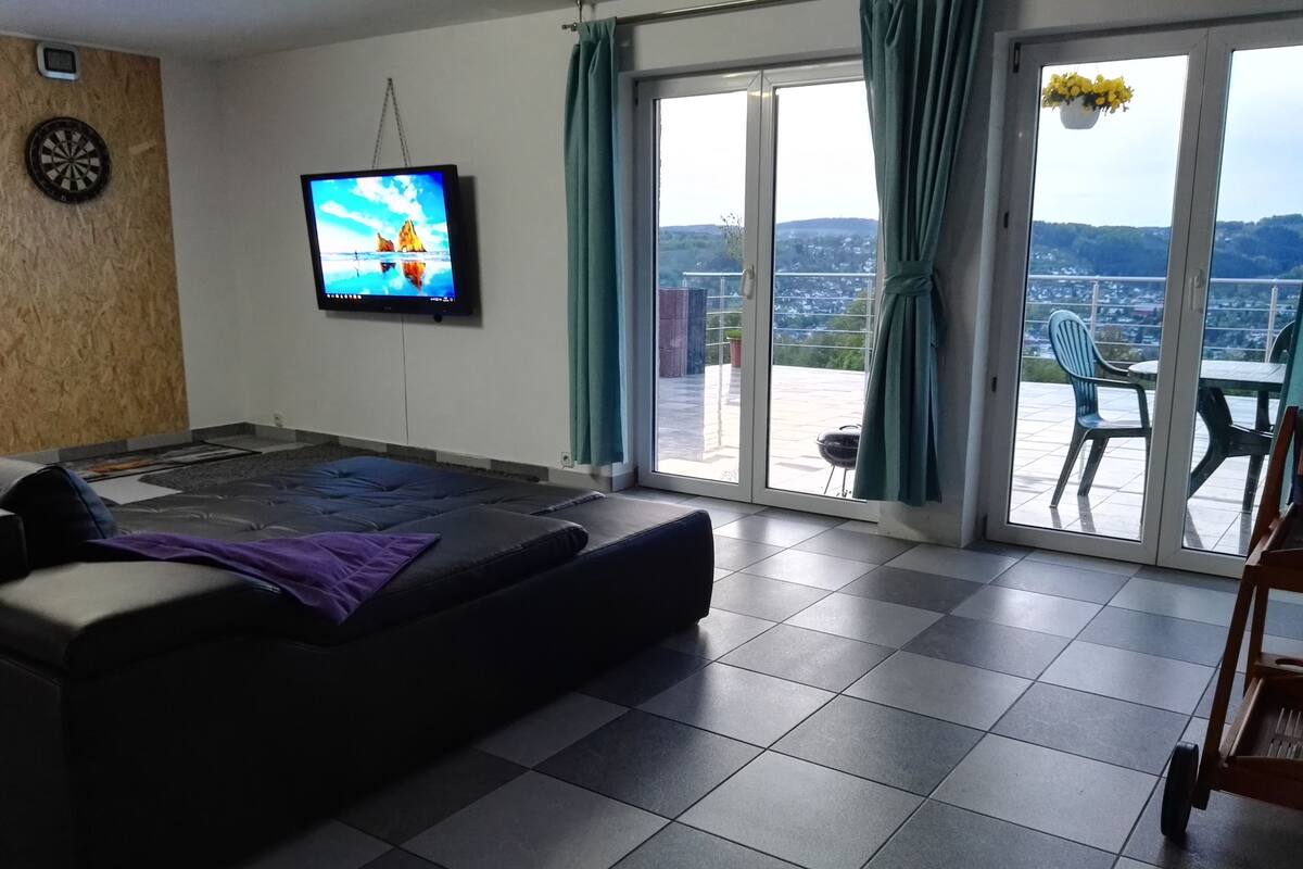 Sala de estar com sofá, TV de tela plana e porta de vidro deslizante para uma varanda com móveis externos.
