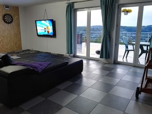 Een woonkamer met een bank, een flatscreen televisie en een schuifdeur naar een balkon met tuinmeubilair.
