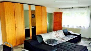 A room with a black sofa, a wooden wardrobe, a small table, and a window with curtains. - Ferienwohnungen nähe Badesee (Bergneustadt)