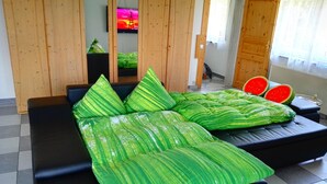 A modern room with a black sofa and green leaf-patterned cushions. A television is mounted on the wall, and a watermelon is visible on the floor. - Ferienwohnungen nähe Badesee (Bergneustadt)