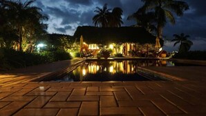 A nighttime view of a resort with a pool, palm trees, and a thatched-roof building.