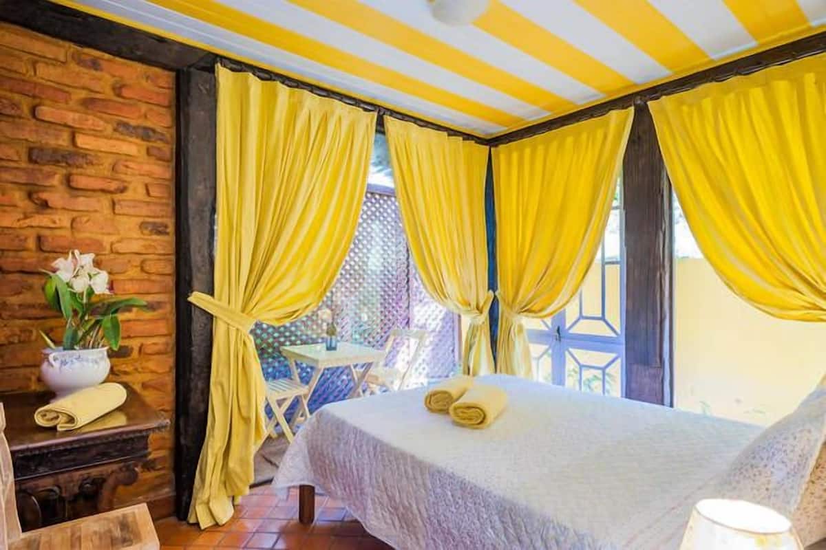 Una habitación con una cama, cortinas amarillas, una mesita con un florero y una pared de ladrillo.