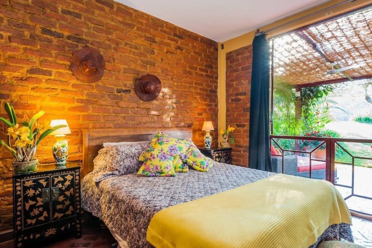 Una habitación con pared de ladrillo, cama con ropa de cama floral, una manta amarilla y vistas a la vegetación exterior.