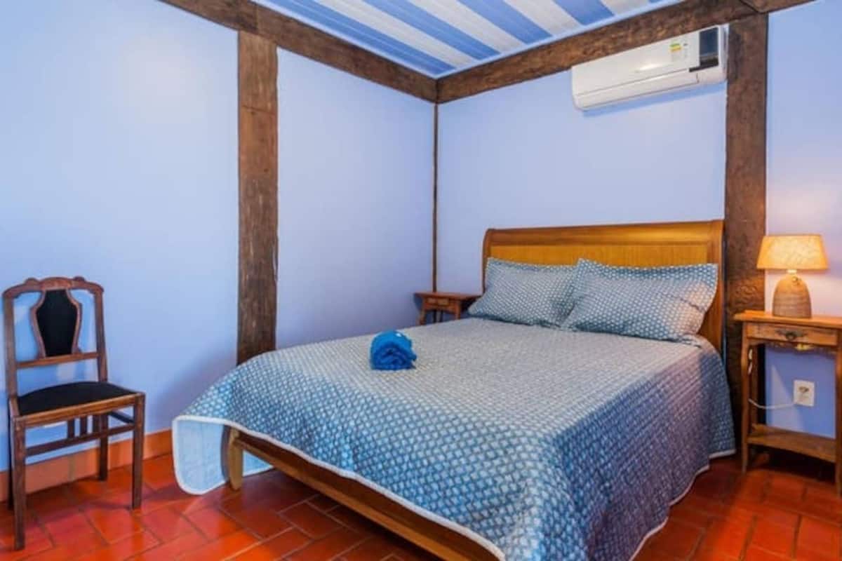 Un dormitorio con cabecera de madera, cama con colcha azul estampada, mesita de noche de madera con lámpara, y una silla.