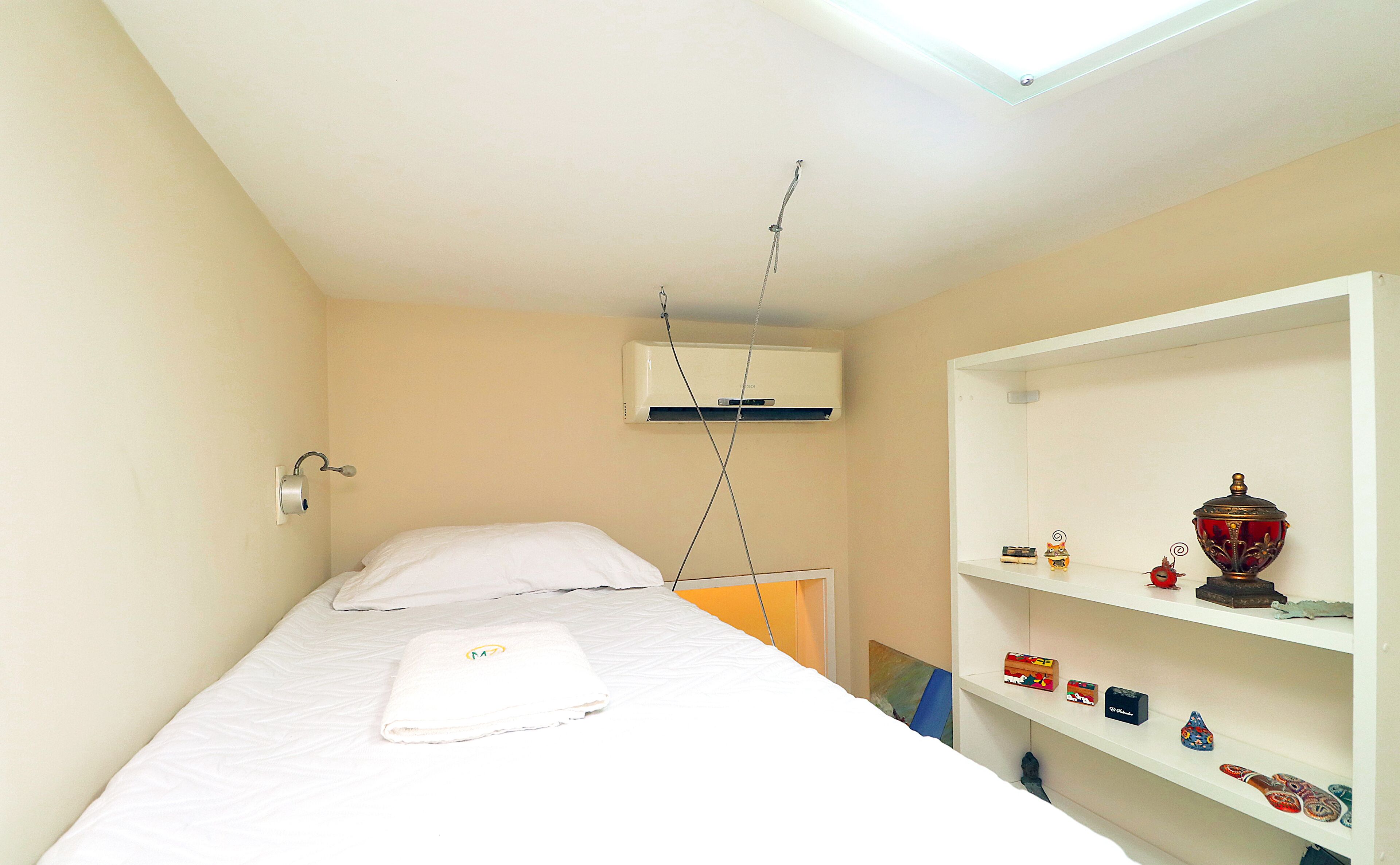 A small, single-bed room with a white bed, a white shelf with decorative items, and a ceiling-mounted air conditioning unit.