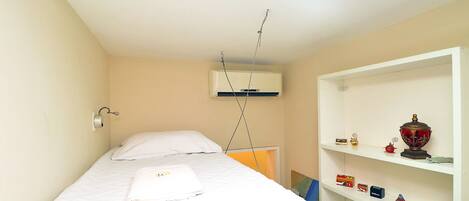 A small, single-bed room with a white bed, a white shelf with decorative items, and a ceiling-mounted air conditioning unit.