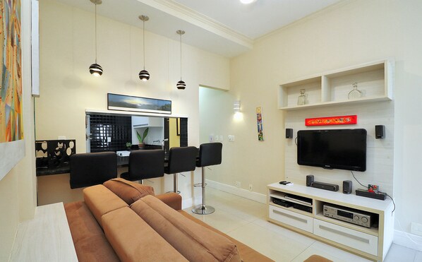 Una sala de estar moderna con un sofá, un televisor de pantalla plana, una mesa de centro y un comedor con sillas, y una cocina al fondo.