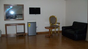 Una habitación con piso de madera, un sofá de cuero negro, una mesita, un sillón con marco dorado, un televisor en la pared y un refrigerador.