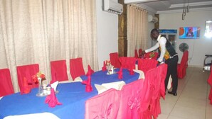 Un área de comedor con mesas cubiertas de manteles azules y fundas rojas para las sillas, una persona ordenando las mesas, y un televisor en la pared.