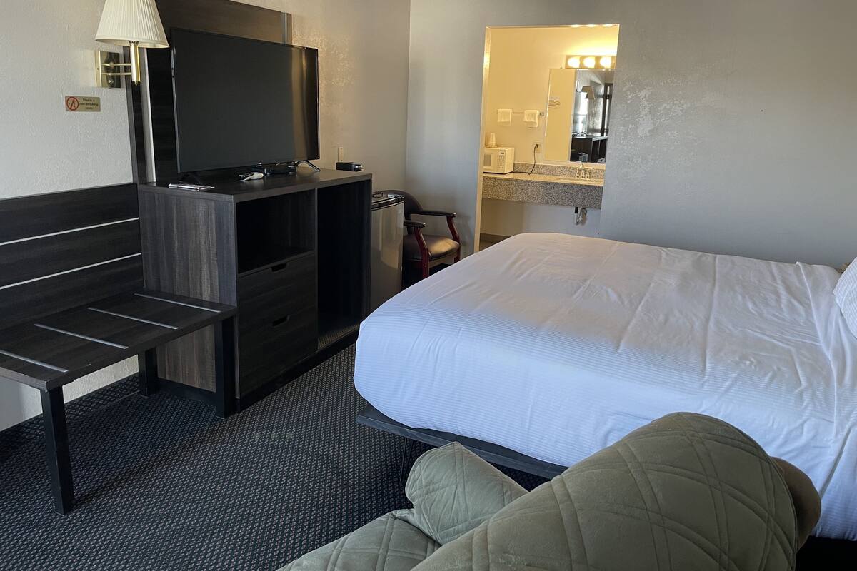 A hotel room with a bed, a desk with a TV, a chair, a small table, and a mirror with lights above the sink.