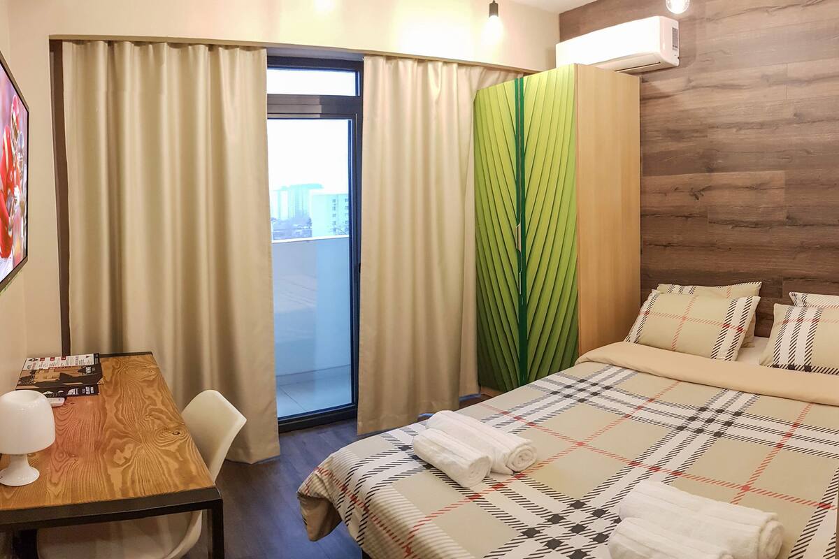 Quarto de hotel com cama, escrivaninha com luminária, janela com cortinas e ar condicionado.