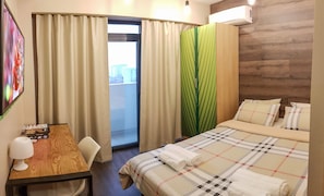 Ein Hotelzimmer mit Bett, Schreibtisch mit Lampe, Fenster mit Vorhängen und einer Klimaanlage.