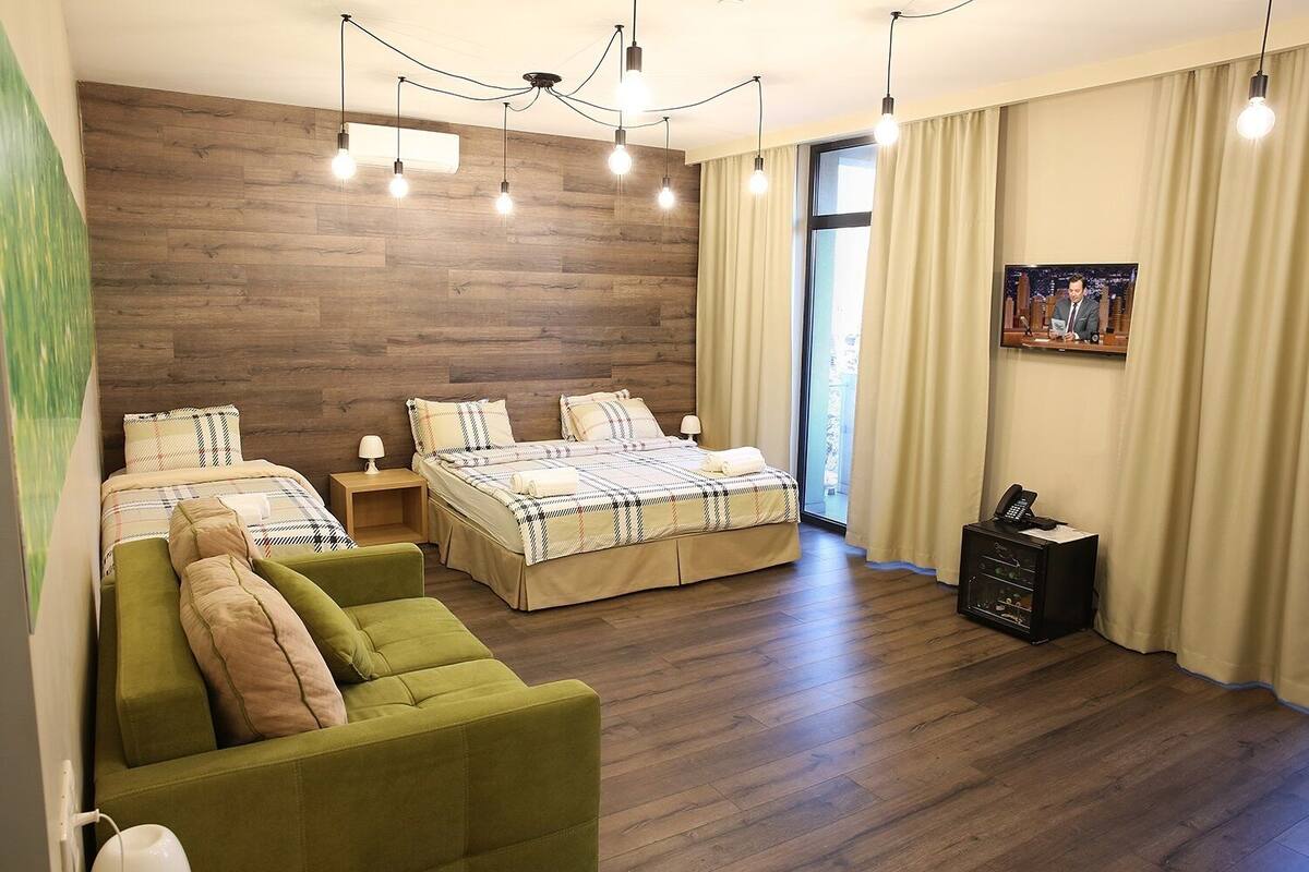 Quarto moderno com parede de madeira, cama com colcha xadrez, sofá verde e escrivaninha com telefone.