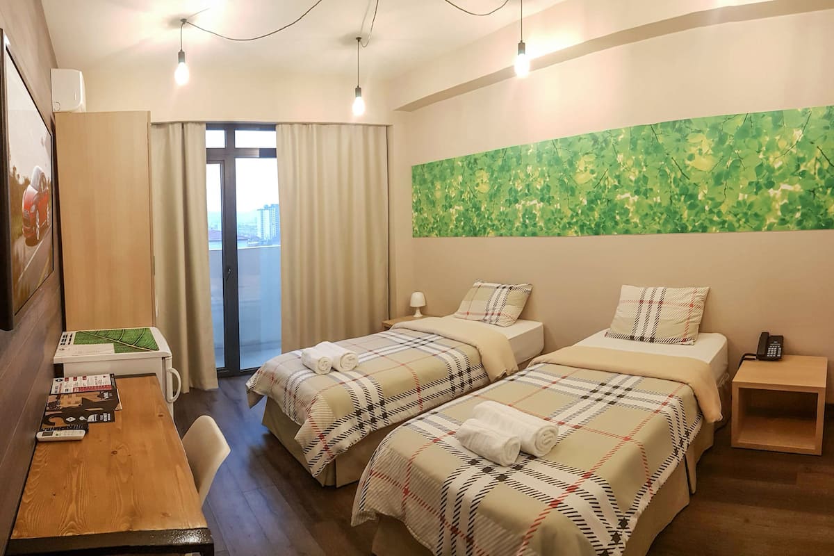 Quarto de hotel com duas camas, uma mesa com cadeira, uma mesinha, telefone e janela com cortinas.