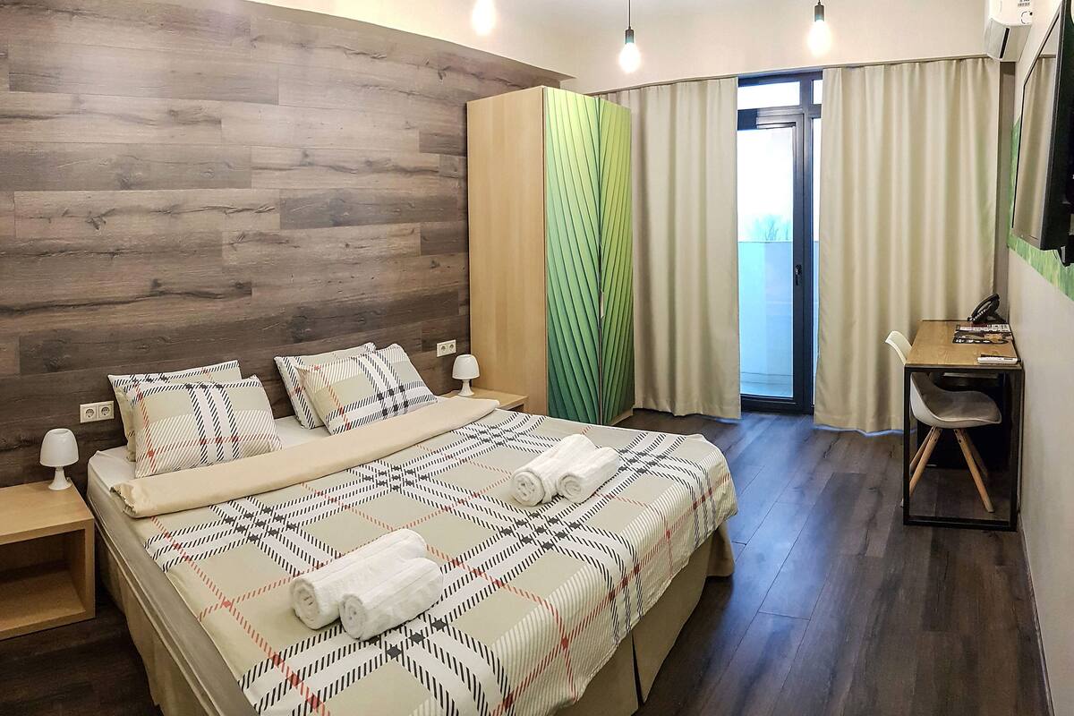 Quarto de hotel com cabeceira de madeira, cama com colcha estampada, duas luminárias de cabeceira, escrivaninha com cadeira e uma janela grande com cortinas.