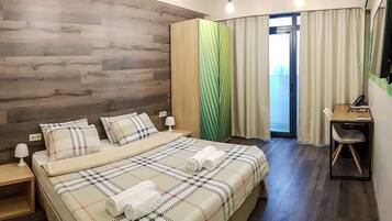 Hotelkamer met een houten hoofdbord, een bed met een beddekleed met patroon, twee bedlampen, een bureau met stoel, en een groot raam met gordijnen.