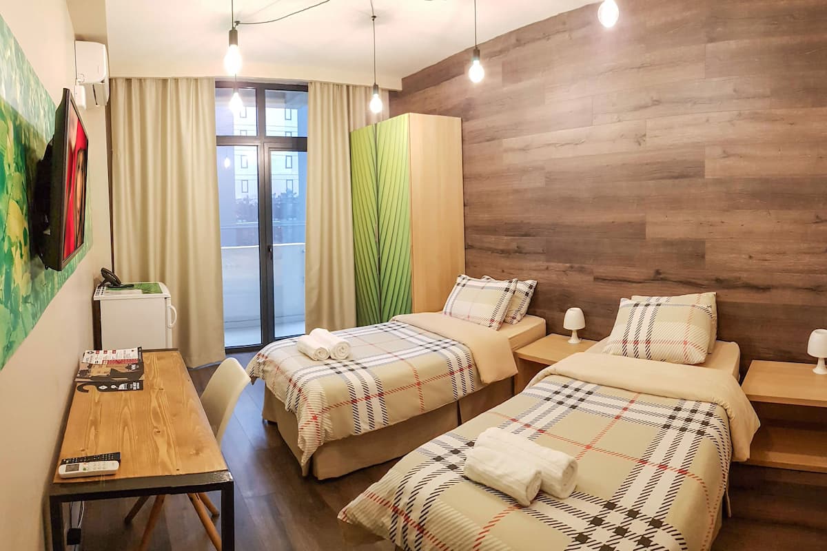 Quarto de hotel com duas camas, uma mesa de madeira, uma televisão, uma escrivaninha e varanda com cortinas.