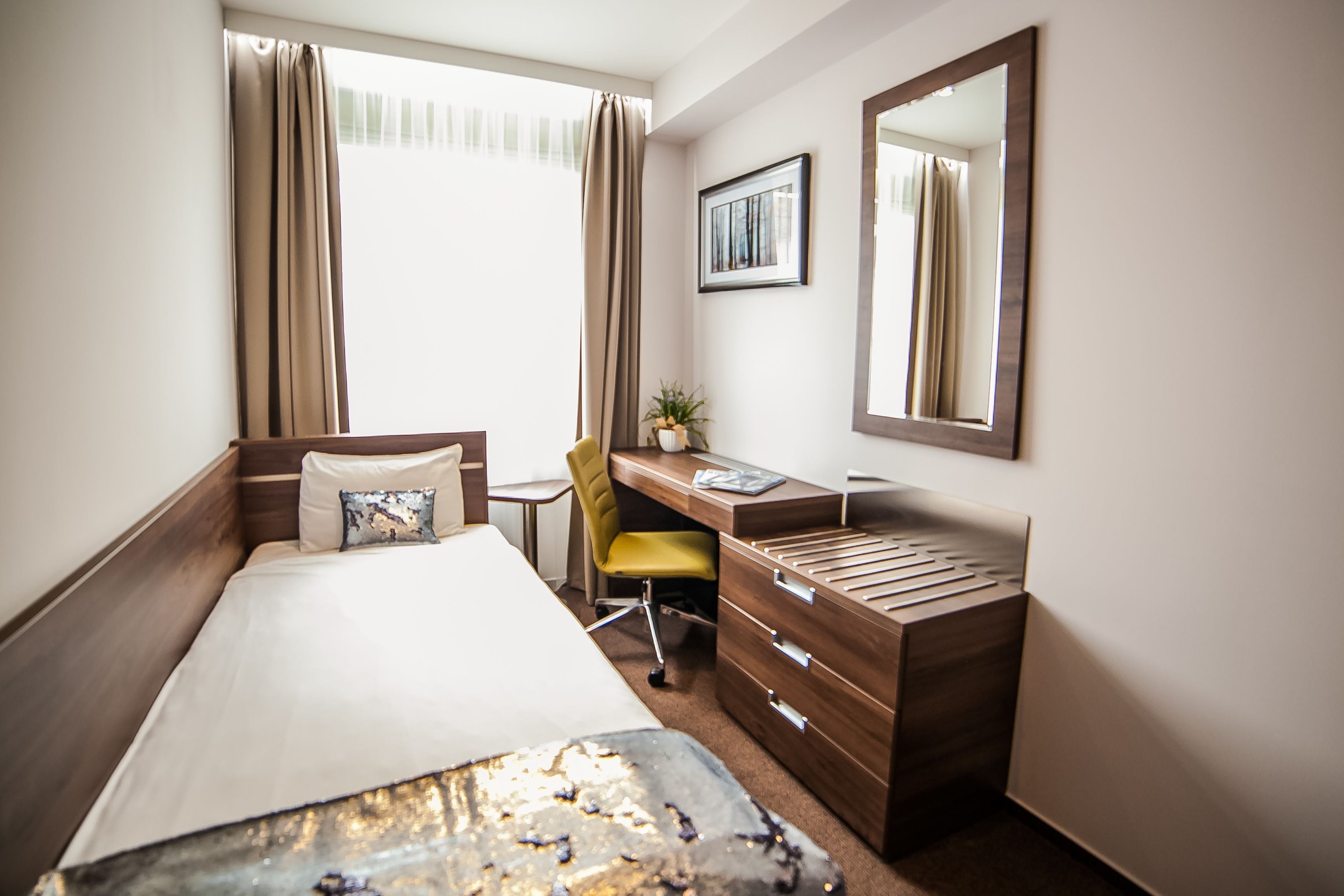 A hotel room with a bed, a desk, a chair, a dresser, a potted plant, and a window with curtains.