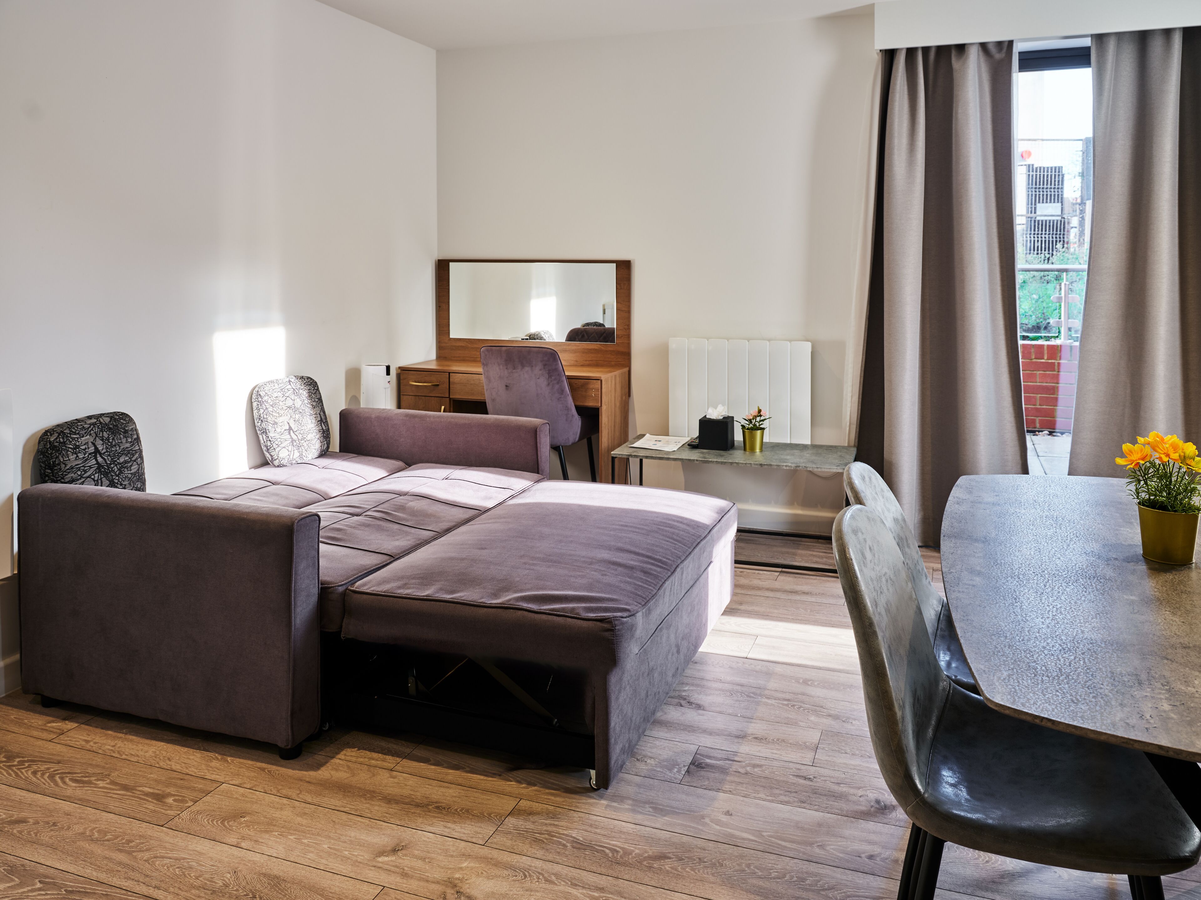 A modern hotel room with a sofa bed, a desk, a chair, and a view of the city through the window.