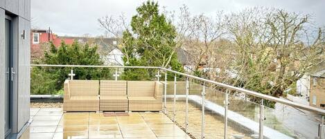 A modern outdoor terrace with a glass balustrade, a wicker sofa, and a stone floor.