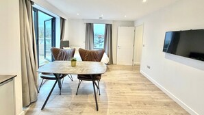 A modern living room with a dining table, two chairs, a TV mounted on the wall, and a large window with curtains. - Hampton Suites Serviced Apartments (Thames Ditton)