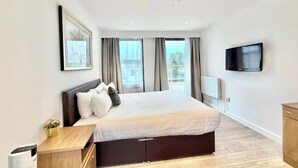 A modern hotel room with a large bed, a TV mounted on the wall, and a window with curtains. - Hampton Suites Serviced Apartments (Thames Ditton)