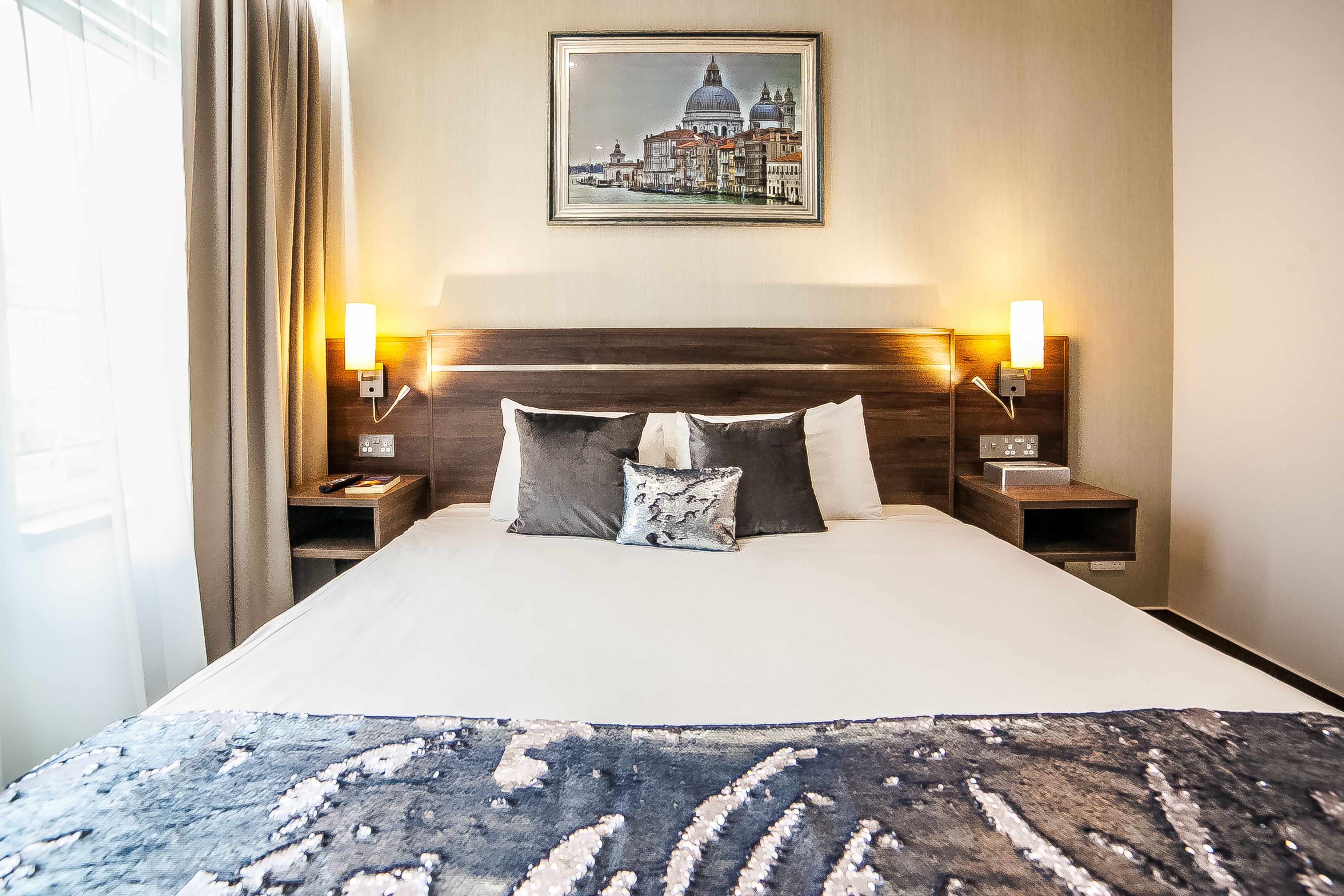 A hotel room with a large bed, two bedside tables with lamps, a framed cityscape picture on the wall, and a window with curtains.