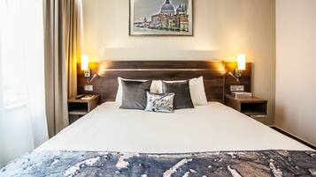 A hotel room with a large bed, two bedside tables with lamps, a framed cityscape picture on the wall, and a window with curtains.