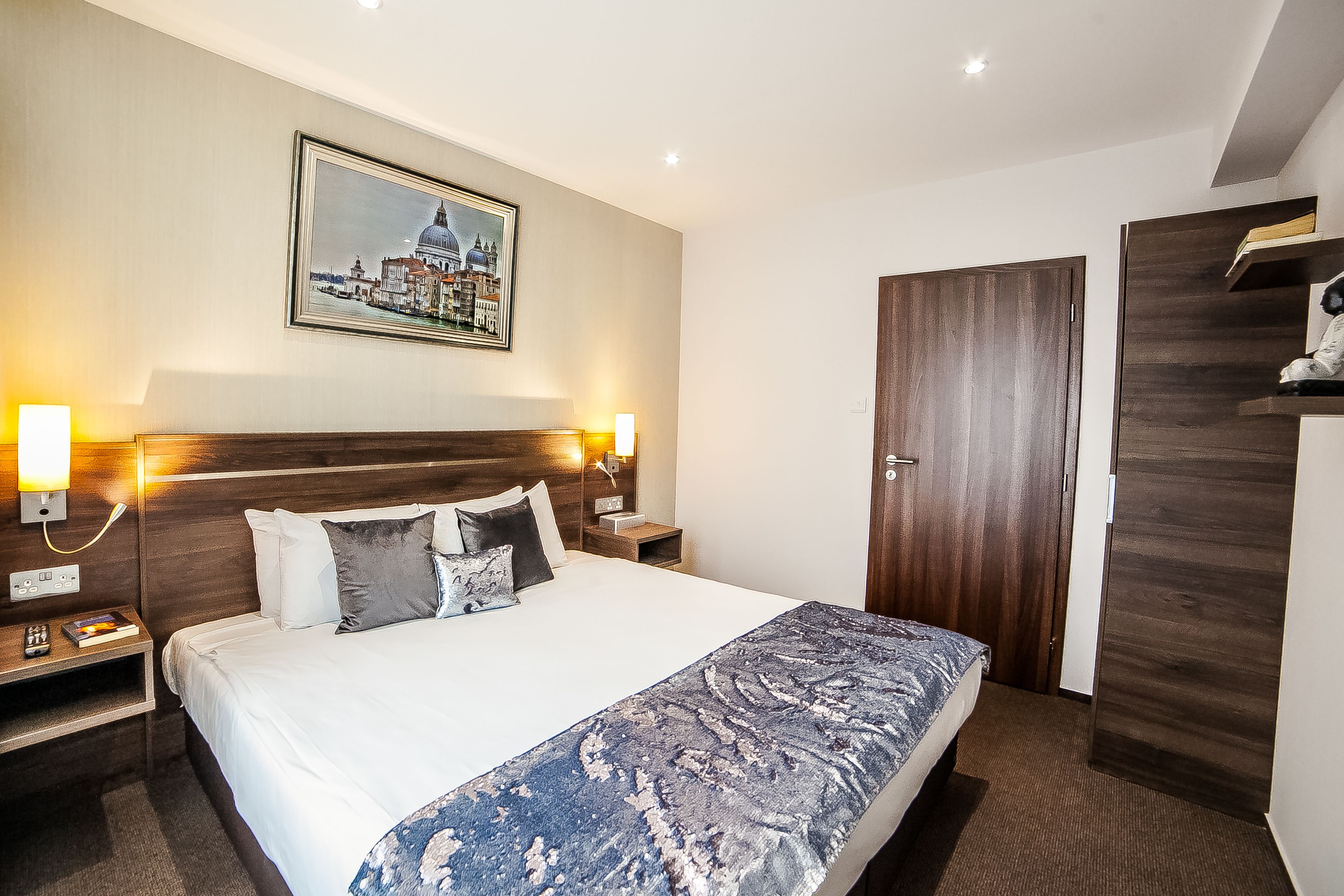 A hotel room with a large bed, wooden headboard, two bedside tables with lamps, a nightstand, a wooden wardrobe, and a framed picture on the wall.