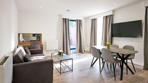 A modern living room with a sofa, coffee table, and dining table with chairs. There is a flat-screen TV mounted on the wall and a window with curtains. - Hampton Suites Serviced Apartments (Thames Ditton)