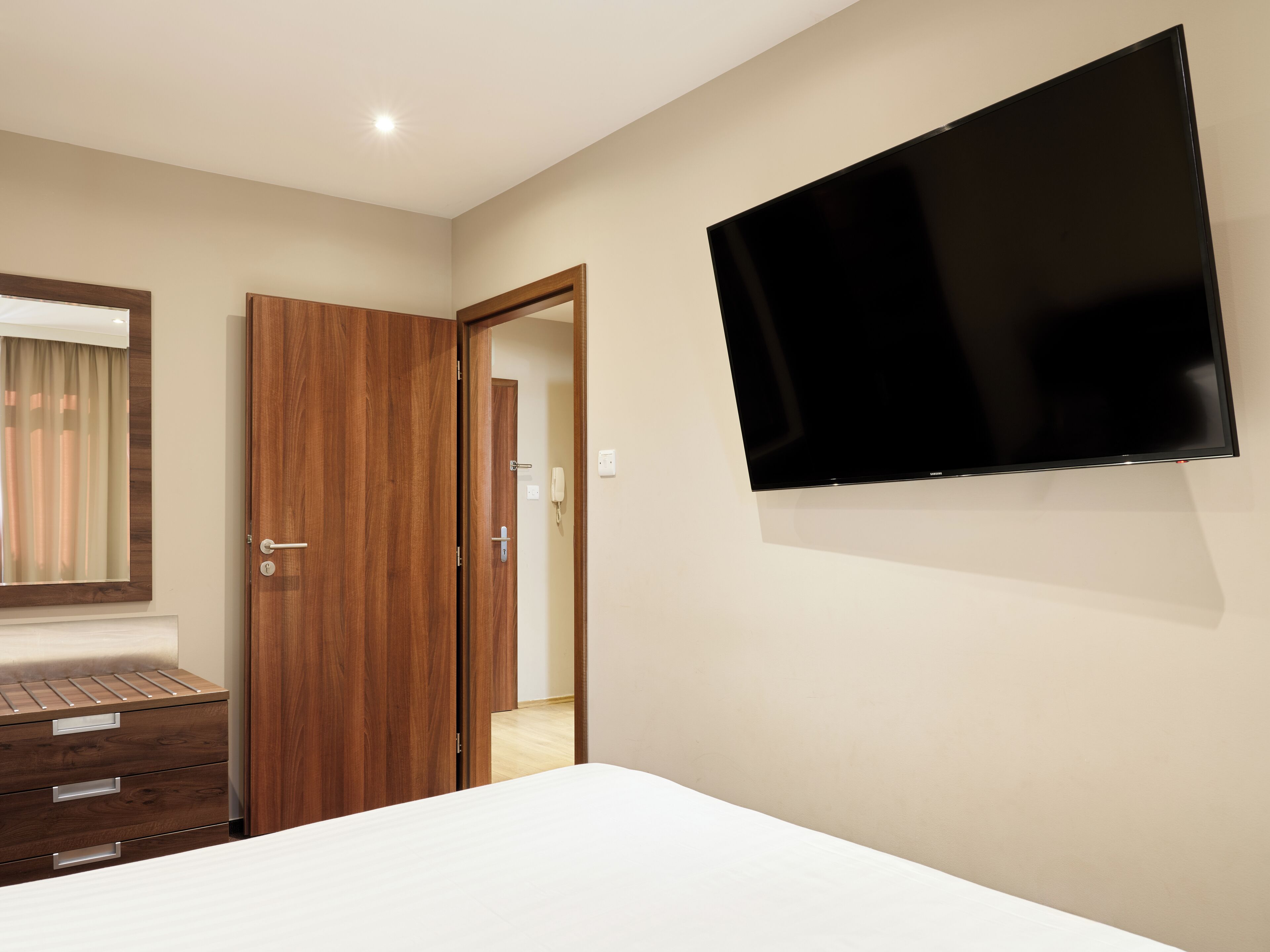 A hotel room with a bed, a wooden wardrobe, a flat-screen TV mounted on the wall, and a door leading to another room.