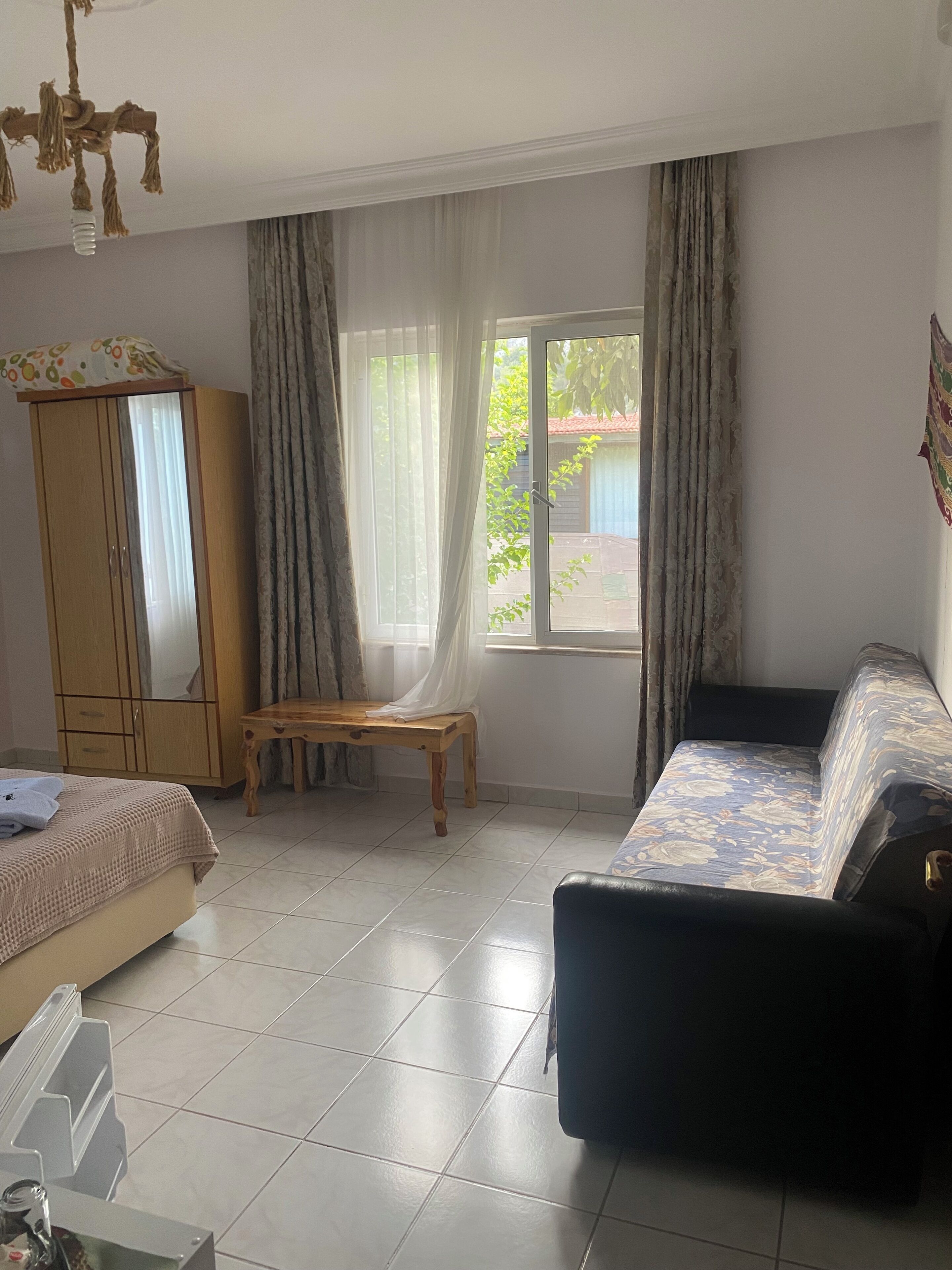 A room with a bed, a sofa, a wooden wardrobe, a table, and a window with curtains.