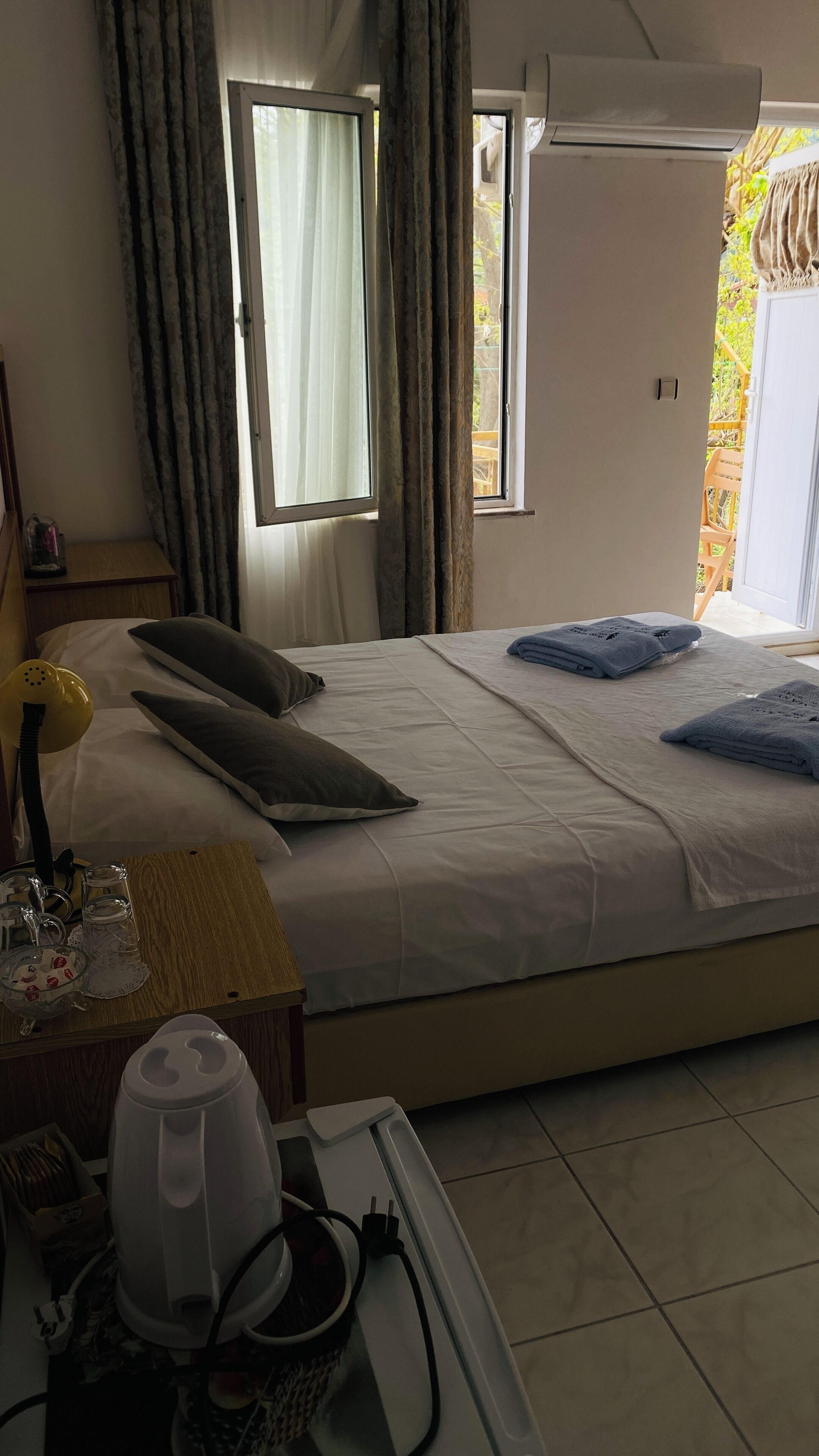 A hotel room with a bed, a bedside table with a lamp and glasses, a window with curtains, and a view of the outdoors.