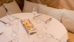 Una mesa redonda para comer con mantel blanco, puesta con copas de vino, cubiertos y una cesta de pan.
