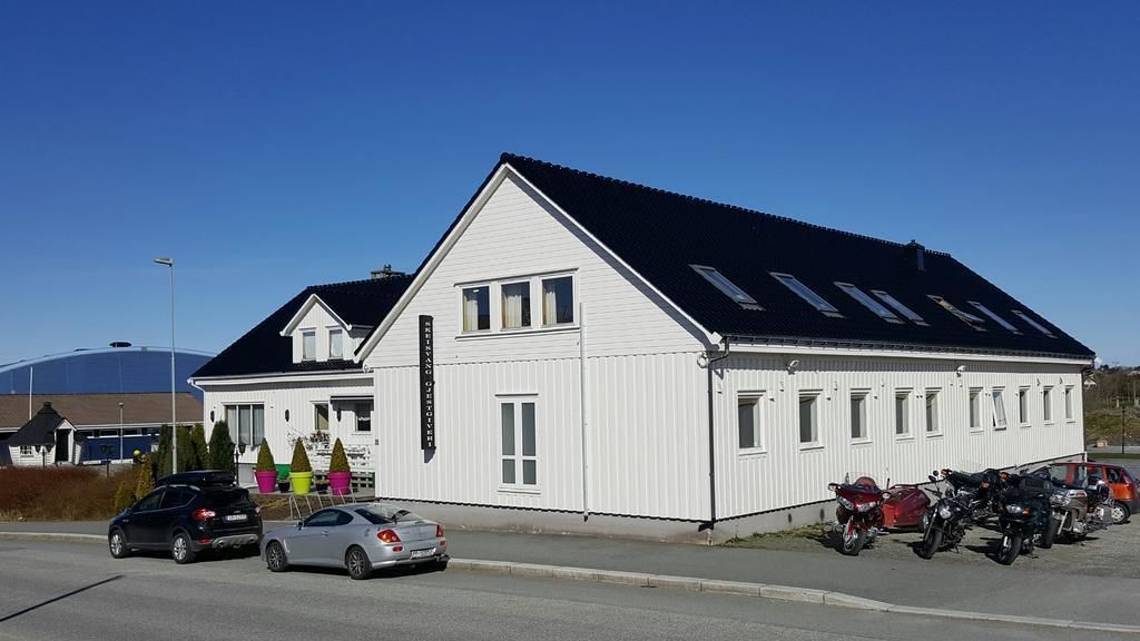 A white building with a black roof, several windows, and a parking area with cars and motorcycles.
