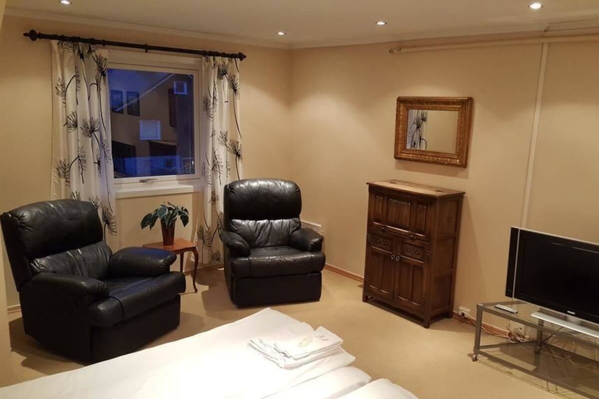 Habitación de hotel con cama, dos sillones de cuero, un armario de madera, televisor sobre un pedestal de vidrio y una ventana con cortinas.