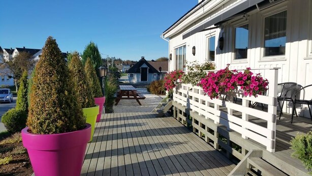Una terraza de madera con una mesa de picnic, plantas en macetas y una cerca blanca adornada con cestas de flores colgantes.