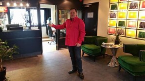 Un hombre con una camisa roja de pie en un vestíbulo con sillones tapizados en verde, un mostrador de recepción y una pared de lámparas estampadas.
