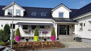 Una casa de dos pisos con techo de tejas oscuras, paredes exteriores blancas y un porche con un banco. Hay tres macetas con plantas en el porche y un pequeño jardín con flores al frente de la casa.